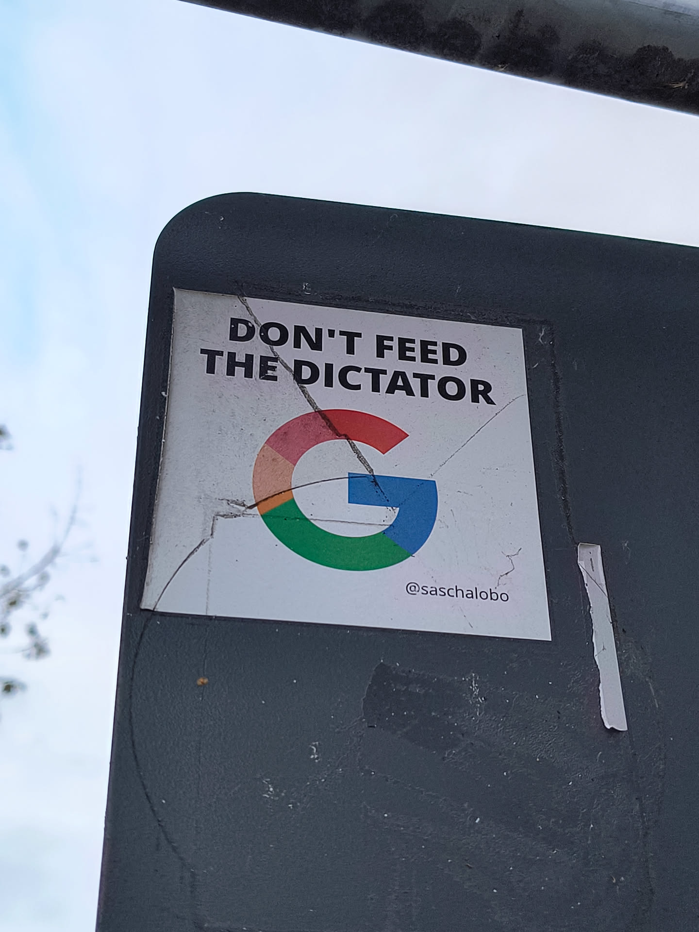 The sticker is rectangular with a white background. At the top, in bold black sans-serif font, it reads 'DON'T FEED THE DICTATOR'. Below this text is a stylized Google 'G' logo, with its characteristic red, yellow, green, and blue segments. The sticker appears to be slightly creased and cracked, suggesting it has been applied outdoors. At the bottom right of the sticker, a social media handle '@saschalobo' is visible in smaller black text.