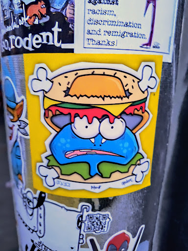 Street sticker A sticker featuring a cartoon monster character eating a burger. The character is blue with big eyes and a grumpy expression. The burger is yellow and dripping with red sauce. The sticker has a yellow background and white bone-like shapes on the sides. There's some text at the bottom that looks like a signature or watermark and numbers 3332.