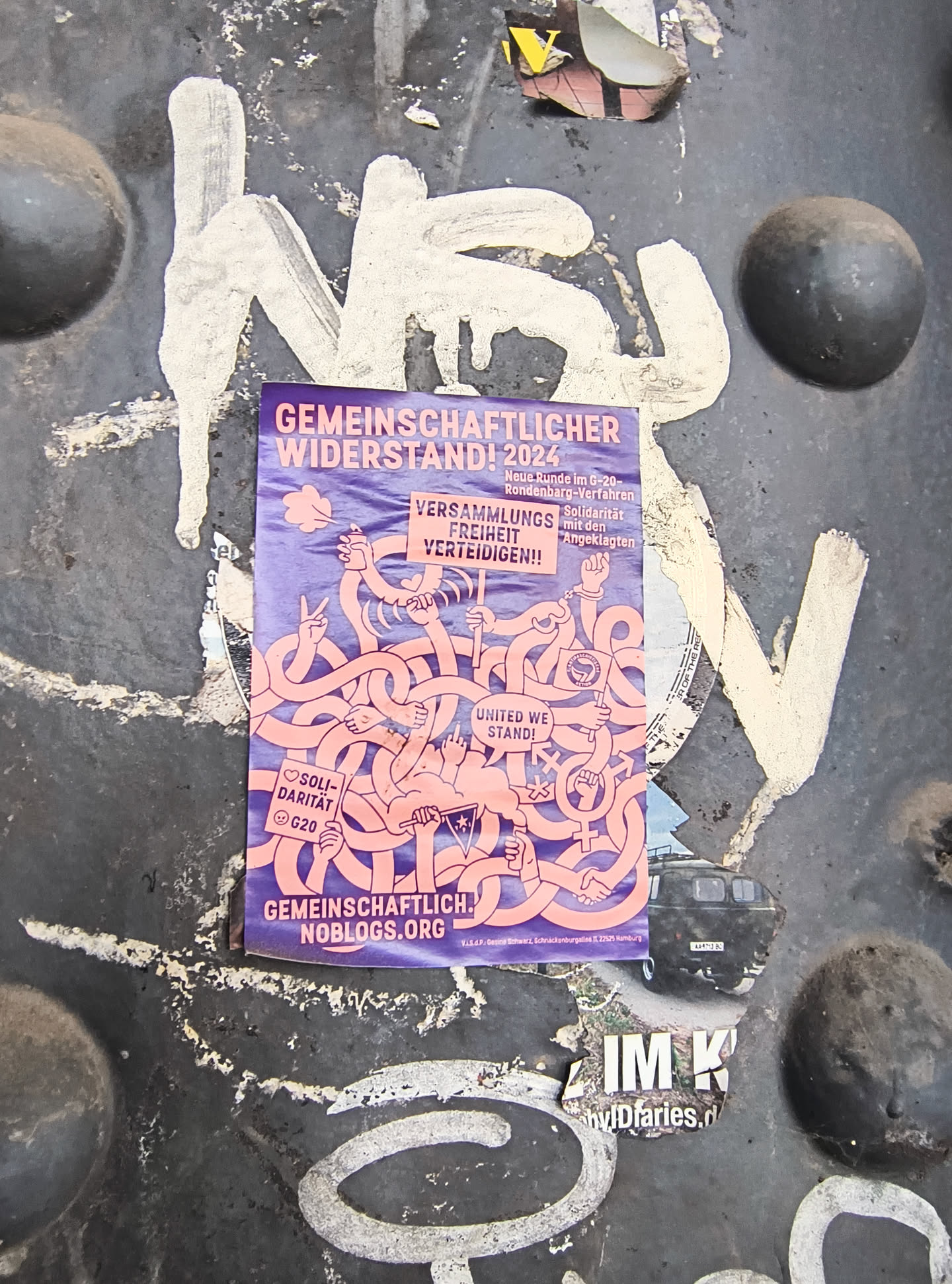 The sticker is primarily purple with pink illustrations. It features a title "GEMEINSCHAFTLICHER WIDERSTAND! 2024" (Community Resistance! 2024) followed by smaller text detailing a 'New round in the G-20Rondenbarg proceedings'. The central graphic depicts a tangled mass of pink arms, many with fists raised or forming peace signs, symbolizing collective action and protest. Several smaller icons are visible, including hearts, a female symbol, a fist, a star within a circle, and an antifa symbol flag. Below the illustration, the text "UNITED WE STAND!" is prominently displayed. Further down, "GEMEINSCHAFTLICH.NOBLOGS.ORG" is printed, indicating a website. The bottom edge of the sticker contains smaller print, likely legal or organizational information.