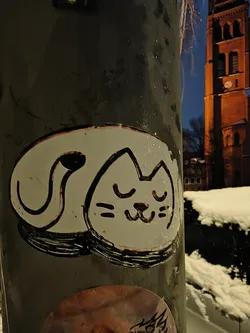 Street sticker The sticker is roughly oval-shaped, featuring a stylized, sleeping cat. The cat is predominantly white with a thick, hand-drawn black outline. Its body is curled up, with its tail forming a distinct loop behind its head. The cat's face shows closed eyes, represented by two short curved lines, and a small, content smile, indicated by two short horizontal lines and a curved line for the mouth. There are subtle reddish or pinkish specks and streaks, particularly around the black outlines and within the white space, suggesting wear or a distressed aesthetic. The overall style is simple, cartoonish, and endearing.