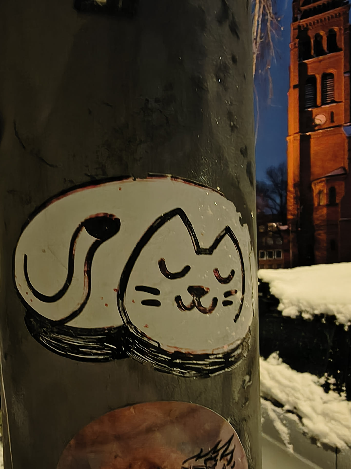 The sticker is roughly oval-shaped, featuring a stylized, sleeping cat. The cat is predominantly white with a thick, hand-drawn black outline. Its body is curled up, with its tail forming a distinct loop behind its head. The cat's face shows closed eyes, represented by two short curved lines, and a small, content smile, indicated by two short horizontal lines and a curved line for the mouth. There are subtle reddish or pinkish specks and streaks, particularly around the black outlines and within the white space, suggesting wear or a distressed aesthetic. The overall style is simple, cartoonish, and endearing.