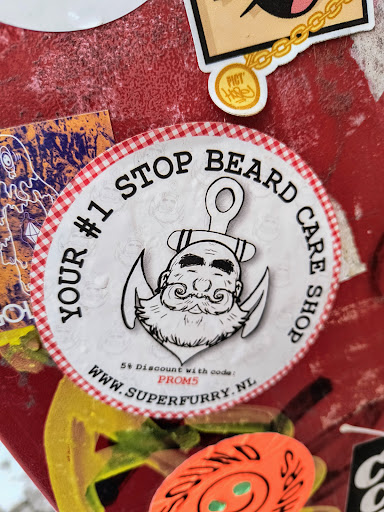 Round sticker with a red and white checkered border featuring an illustration of a bearded man's head with an anchor, along with the text "Your #1 Stop Beard Care Shop", a website address, and a discount code. 