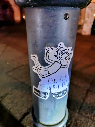 The sticker on the pole depicts a hand-drawn cartoon character in mid-air. The character has distinct features, including oversized eyes. At the bottom is the text 'DELLA GRAPPA' and the url 'www.dellagrappa.de'.