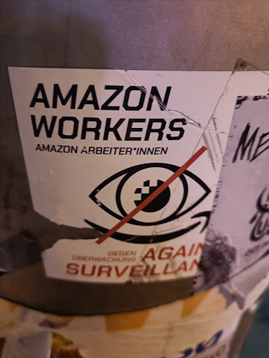 A sticker protesting Amazon and surveillance. The sticker is partially damaged and has text in both English and German.  The main text says "AMAZON WORKERS" and "AMAZON ARBEITER*INNEN" (Amazon workers in German, using gender-inclusive language).  There is also a graphic of an eye with a red line across it, symbolizing the rejection of surveillance, and smaller text that says "GEGEN ÜBERWACHUNG AGAIN SURVEILLANCE".