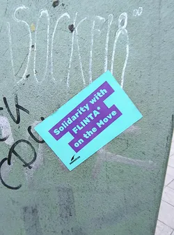 Street sticker A rectangular sticker with a bright turquoise background and bold purple text. The text reads 'Solidarity with FLINTA* on the Move'. Below the main text, in smaller font and a black silhouette of a bird, is the word 'SRBDRUCKE'. The sticker appears to be placed on a textured, greenish-gray surface that has other graffiti markings on it.