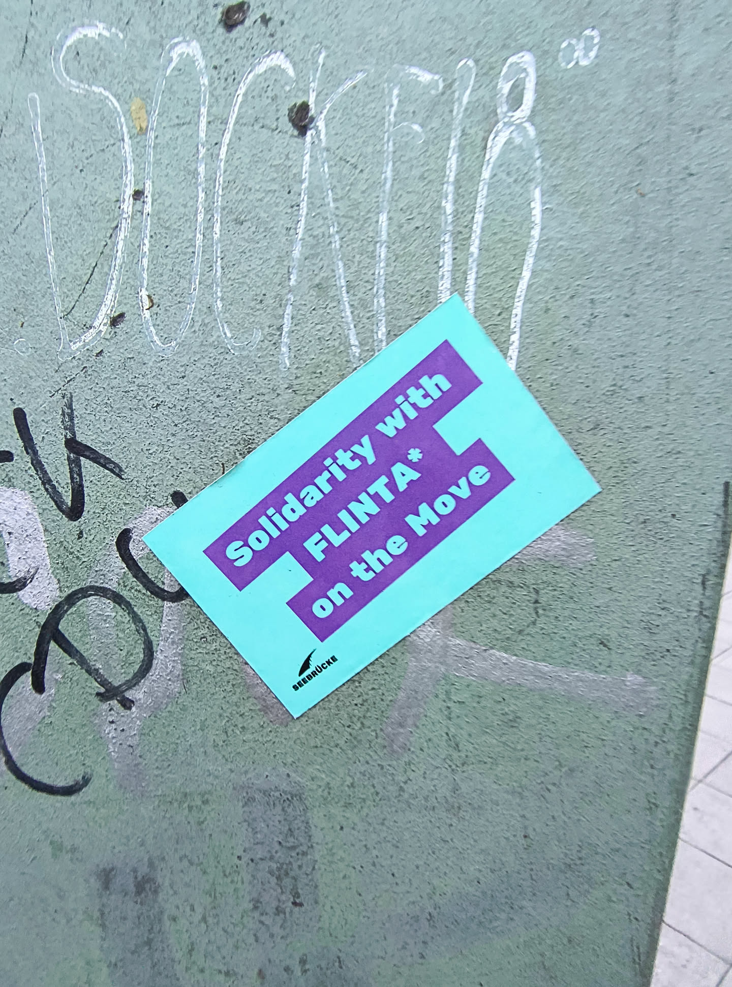 A rectangular sticker with a bright turquoise background and bold purple text. The text reads 'Solidarity with FLINTA* on the Move'. Below the main text, in smaller font and a black silhouette of a bird, is the word 'SRBDRUCKE'. The sticker appears to be placed on a textured, greenish-gray surface that has other graffiti markings on it.