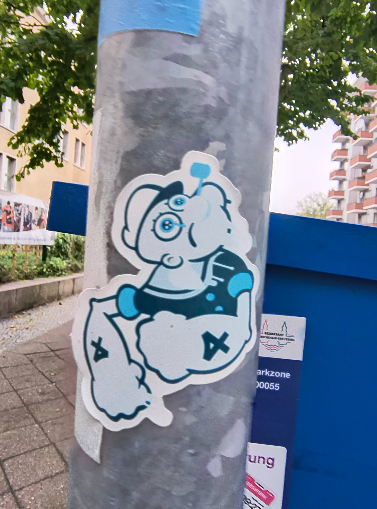 Street sticker The sticker features a cartoon character resembling a Smurf, depicted with a muscular physique. The character has large, round blue eyes with glasses, a white hat, and is wearing a dark blue vest with light blue accents. The character's arms are flexed, and its legs are bent. The sticker is predominantly white with blue and dark blue outlines and details. There are also two 'X' symbols in black on the character's legs.