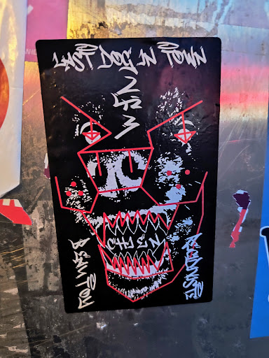 Street sticker A black sticker shows an abstract depiction of a dog's face with red geometric lines and jagged white teeth. The text "LAST DOG IN TOWN" is at the top, with illegible artist signature on the bottom.