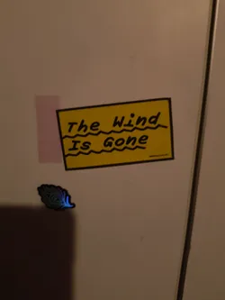 Street sticker The central sticker is a yellow rectangle with a thick black border. Inside, the text 'The Wind' is written on the top line, followed by a jagged, wavy line. Below that, 'Is Gone' is written on the second line, also with a jagged, wavy line beneath it. The text is in a handwritten, slightly whimsical font. To the left and slightly below the yellow sticker is a smaller, distinct sticker depicting a stylized feather or leaf-like shape. This smaller sticker has a dark outline and is filled with a gradient of blues and greens, with a bright blue glow emanating from its tip, suggesting an iridescent or luminous quality.