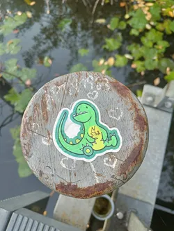 A green dinosaur sticker is attached to a rusty grey wooden post by a body of water. The sticker shows a cartoonish dinosaur sitting down. There are some writings and drawings on the wooden post around the sticker. 