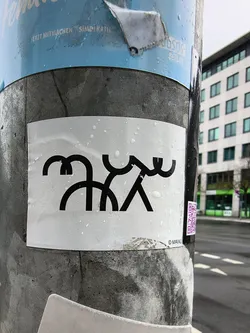 Street sticker A rectangular white sticker, slightly creased and covered in water droplets, adheres to a grey pole. Its central design is a striking black line art illustration. The artwork depicts two highly stylized, abstract figures. The left figure is composed of rounded, interconnected black lines, resembling a head and body with arms. The right figure features more angular lines, forming a body and limbs, with a wavy top element. The two figures appear to be linked or interacting in the center. In the bottom right corner of the sticker, a small copyright symbol is followed by the name 'MAIXU' in black text.