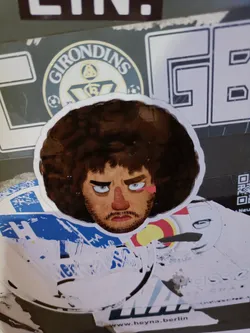 Street sticker Close-up of a round sticker featuring a cartoon-like illustration of a man with a voluminous, dark brown afro. His facial features are exaggerated, with large, expressive eyes and a serious expression.  The art style is reminiscent of a painted portrait, with visible brushstrokes. The sticker appears to be affixed to a wall or surface with other stickers and markings around it.