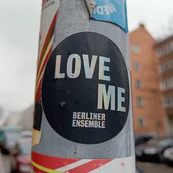 Street sticker A round black and white sticker with the text "LOVE ME" in a holographic foil and "BERLINER ENSEMBLE" underneath. The sticker is affixed to a pole, with various other stickers visible on the surface.