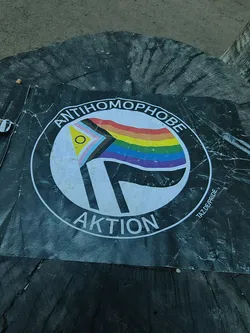 Street sticker Circular sticker on a black background featuring a Progress Pride flag and a black flag, styled after the Antifascist Action logo. Text reads 'ANTIHOMOPHOBE AKTION' with 'TAZ.DE/PRIDE' on the side.