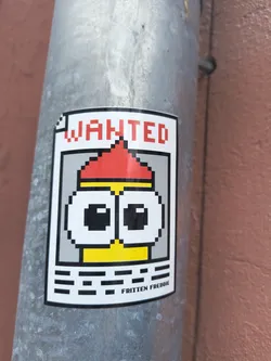Street sticker A rectangular sticker designed as a 'WANTED' poster, featuring a pixel art character. The sticker has a white background with a thick black border. At the top, the word 'WANTED' is written in bold, red, pixelated capital letters. Below this, a central character is depicted in a pixelated style, resembling a Minion-like figure. It has a yellow body/face, large white eyes with black pupils, and a red pointed hat or hair, separated by a thin yellow band. Below the character, there are several abstract horizontal black pixelated lines, suggesting a space for information. At the very bottom, centered, the name 'FRITTEN FREDDIE' is written in black, pixelated letters. The sticker is adhered to a textured, grey metallic pole, showing slight wear and a subtle curve.