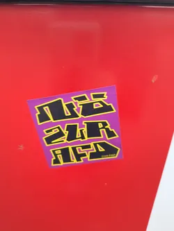 Street sticker The sticker is square-ish with slightly rounded corners and is purple in color. It features bold, blocky lettering in black with a thick yellow outline. The text appears to be arranged in three lines. The top line has abstract shapes that resemble letters. The second line clearly reads '2UR'. The third line reads 'AFD'. In the bottom right corner of the sticker, there is a small social media handle '@noe.digga'.