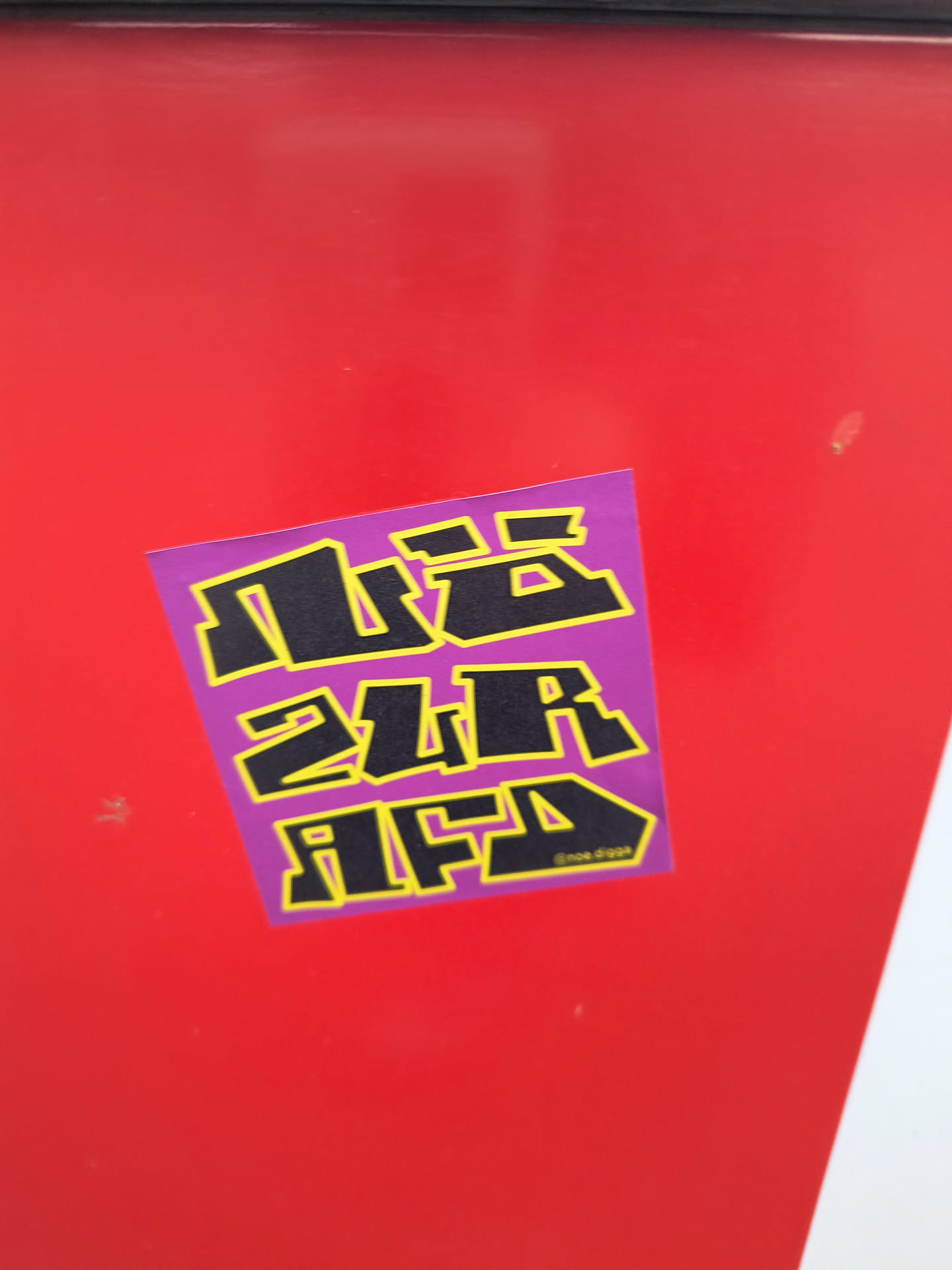 The sticker is square-ish with slightly rounded corners and is purple in color. It features bold, blocky lettering in black with a thick yellow outline. The text appears to be arranged in three lines. The top line has abstract shapes that resemble letters. The second line clearly reads '2UR'. The third line reads 'AFD'. In the bottom right corner of the sticker, there is a small social media handle '@noe.digga'.