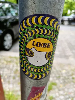 Street sticker A round sticker featuring a cartoon face with the word "Liebe" (German for Love) on its forehead. The face is surrounded by a mesmerizing pattern of yellow and purple crescent shapes on a green background, creating an optical illusion effect. The sticker is affixed to a metal pole.