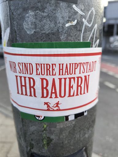 A sticker on a pole with a bold text in German that translates to "We are your capital, your farmers." It also features a small graphic of two figures seemingly running or playing. The sticker has a green stripe at the top and bottom.