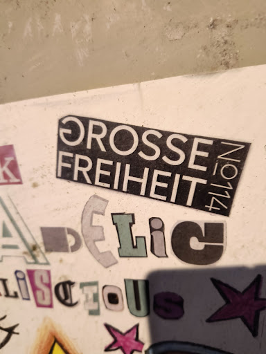 Street sticker A black rectangle sticker with the words "GROSSE FREIHEIT No. 114" in white, pasted among other stickers on a white surface. The other stickers are various letters cut out from magazines, giving a collage effect.