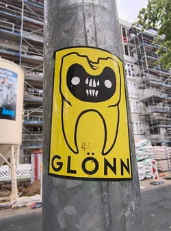 Street sticker The sticker is predominantly yellow with a thick black outline. At its center is a stylized, anthropomorphic tooth character. The character has a black face with two large white circles for eyes and a wide mouth filled with sharp, white, pointed teeth. Above the eyes, it has two small black indentations that could be interpreted as eyebrows or horns. The body of the character is a rounded, bulbous shape, also outlined in black. Below the character, in bold black sans-serif font, is the word 'GLÖNN'. The sticker appears to be slightly worn with a few small rust-colored marks on the yellow background.