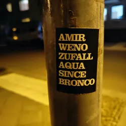 Street sticker A black sticker on a metal pole with white text reading: AMIR WENO ZUFALL AQUA SINCE BRONCO. The words are stacked vertically.