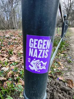 Street sticker A rectangular sticker, predominantly vibrant purple with white text and imagery. The top half features the bold, sans-serif text "GEGEN NAZIS" (German for "Against Nazis"). Below the text, a stylized white graphic depicts a hand breaking a swastika symbol. The sticker shows minor wear, with a slight peel visible at its top-left corner, and is affixed to a dark, possibly black or dark grey, metal pole.