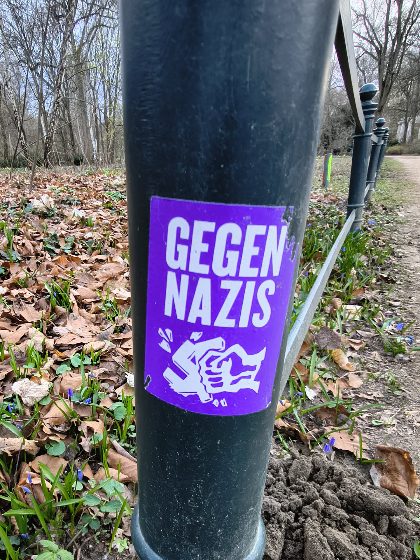 A rectangular sticker, predominantly vibrant purple with white text and imagery. The top half features the bold, sans-serif text "GEGEN NAZIS" (German for "Against Nazis"). Below the text, a stylized white graphic depicts a hand breaking a swastika symbol. The sticker shows minor wear, with a slight peel visible at its top-left corner, and is affixed to a dark, possibly black or dark grey, metal pole.