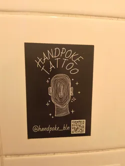 Street sticker A rectangular black sticker featuring white line art. At the top, the words 'HANDPOKE TATTOO' are written in an arched, handwritten style. Below this, a central illustration depicts the outline of a human head, filled with intricate, organic, concentric lines resembling a fingerprint or brain contours. Small white stars and crosses are scattered around the head, suggesting a subtle sparkle or aura. At the bottom left, the social media handle '@handpoke_bln' is written in a cursive font. To the bottom right, a square QR code is prominently displayed.
