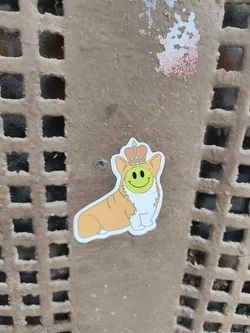 Street sticker The sticker features a stylized depiction of a corgi with its body facing left and its head turned slightly to the right. The corgi is primarily orange with white fur around its neck and chest, and white paws. It is wearing a golden crown with red and blue accents on its head. Its face is a bright green smiley face with black dot eyes and a black curved mouth. The sticker has a white border.
