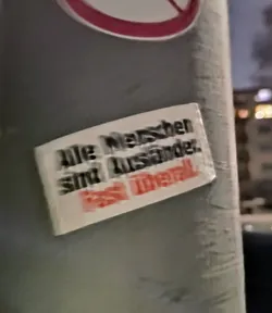 Street sticker A sticker with the text "Alle Menschen sind wunderbar. Es ist Moral." which translates to "All people are wonderful. It's morals." in German. The sticker is attached to a gray pole.