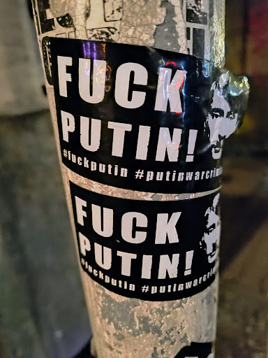 Two black and white stickers plastered on a pole. The stickers read "FUCK PUTIN!" with hashtags #fuckputin and #putinwarcriminal. There's also a cutout of Putin's head on the right side of the sticker.