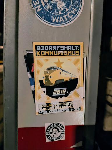 A sticker featuring a retro-style illustration of a train with the text "Bedarfshalt: Kommunismus" (Demand stop: Communism) and the year 2019. The design is reminiscent of socialist propaganda posters.