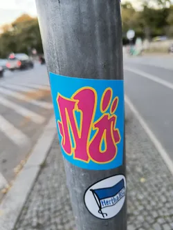 A vibrant sticker featuring a stylized pink 'N' on a bright blue background. The sticker is affixed to a metal post. No QR code or social media links found.