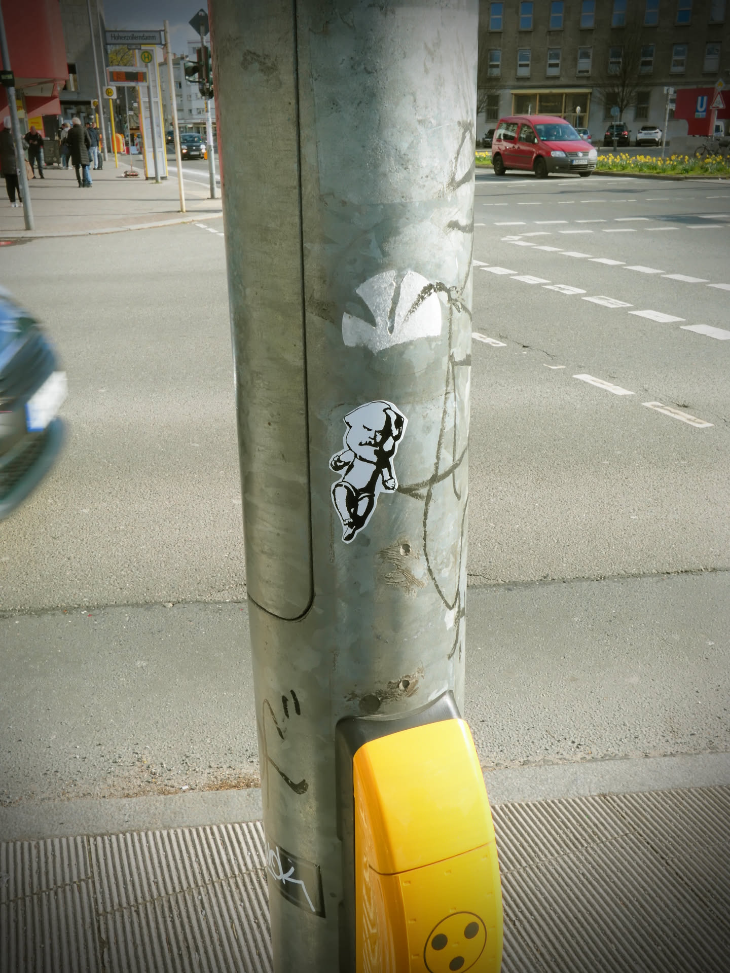 A monochrome sticker featuring a stylized, somewhat chubby baby or infant figure. The figure is depicted in a dynamic pose, possibly running or floating, with one leg bent and the other extended. One arm is raised, forming a clenched fist, suggesting determination or defiance. The face has a determined or slightly angry expression, with a furrowed brow. The sticker uses bold black outlines against a white or light grey interior, giving it a stark, illustrative appearance. It is affixed to a textured, metallic pole.