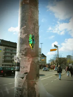 Street sticker The sticker depicts a stylized, somewhat chubby baby or cherubic figure rendered in an iridescent or holographic material. Its colors shift dynamically between vibrant greens, blues, and yellows, creating a shimmering effect. The figure is shown in a crawling or sitting pose, with its head slightly tilted and a distinct, somewhat serious or grumpy expression. A dark outline defines the figure against the bright, shifting internal colors. It is affixed to a weathered metal pole.