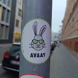 Street sticker A round sticker featuring a cartoon rabbit wearing sunglasses. The text 'AVAAAY' is written below the rabbit.  The sticker is attached to a metal pole on a city street.