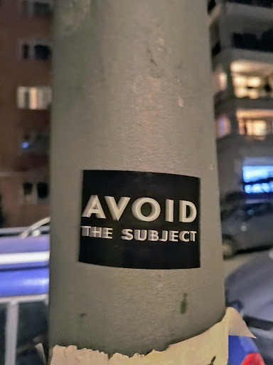 A black and white sticker with the text "AVOID THE SUBJECT" in a sans-serif font. The sticker is applied to a gray pole, with some of the adhesive showing at the bottom. The background is slightly blurred and shows a city street at night.