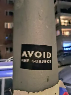 Street sticker A black and white sticker with the text "AVOID THE SUBJECT" in a sans-serif font. The sticker is applied to a gray pole, with some of the adhesive showing at the bottom. The background is slightly blurred and shows a city street at night.