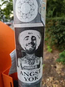 Black and white sticker of a man with a third eye on his forehead, wearing a hat. The sticker has the text "Angst Yok" at the bottom and "RELIEWS" in a small rectangular box on the man's chest.