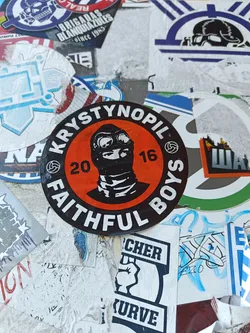 Street sticker A round sticker with a black and orange color scheme. It has an illustration of a person wearing a balaclava and goggles in the center. The text "Krystynopil" and "Faithful Boys" are written around the illustration, with the numbers "20" and "16" flanking the illustration. The style is reminiscent of a logo or emblem.