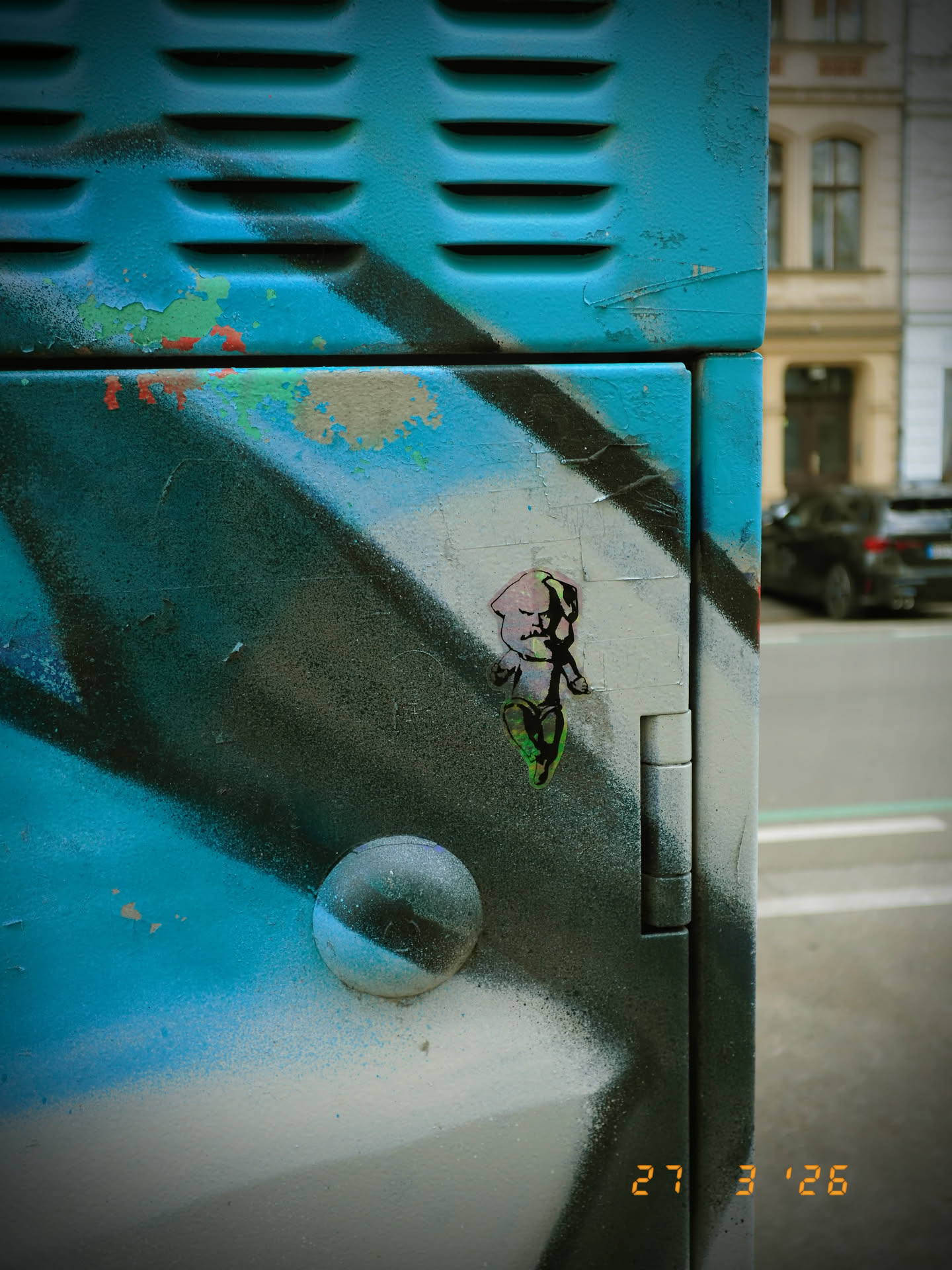 The sticker features a stylized, cartoonish figure resembling a baby or cherub, rendered with a distinct, somewhat edgy aesthetic. The figure has a large, round head with a light pink or flesh-toned complexion, outlined in dark black. Its hair is a striking, bright neon green, giving it a punk-rock appearance. The eyes are small black dots, and the mouth is a simple, dark line. The body is small and disproportionate to the head, with short limbs. The legs and feet area also show hints of the same vibrant green. The entire figure is outlined in black, creating a bold contrast against the lighter background of the sticker and the painted surface it adheres to. The sticker itself has a slightly irregular, hand-cut appearance.