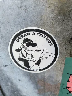 Street sticker The sticker is circular and features a black and white cartoon image of a dog with sunglasses giving a pointing gesture. The words "Urban Attitude" are printed around the top edge of the circle. The sticker is on a metallic surface.