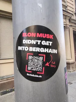 Street sticker This is a round black sticker attached to a metal pole. The text says, in all caps, "Elon Musk DIDN'T GET INTO BERGHAIN" with a QR code and the text "Full Story" next to it, as well as "BerlinGuide.de".