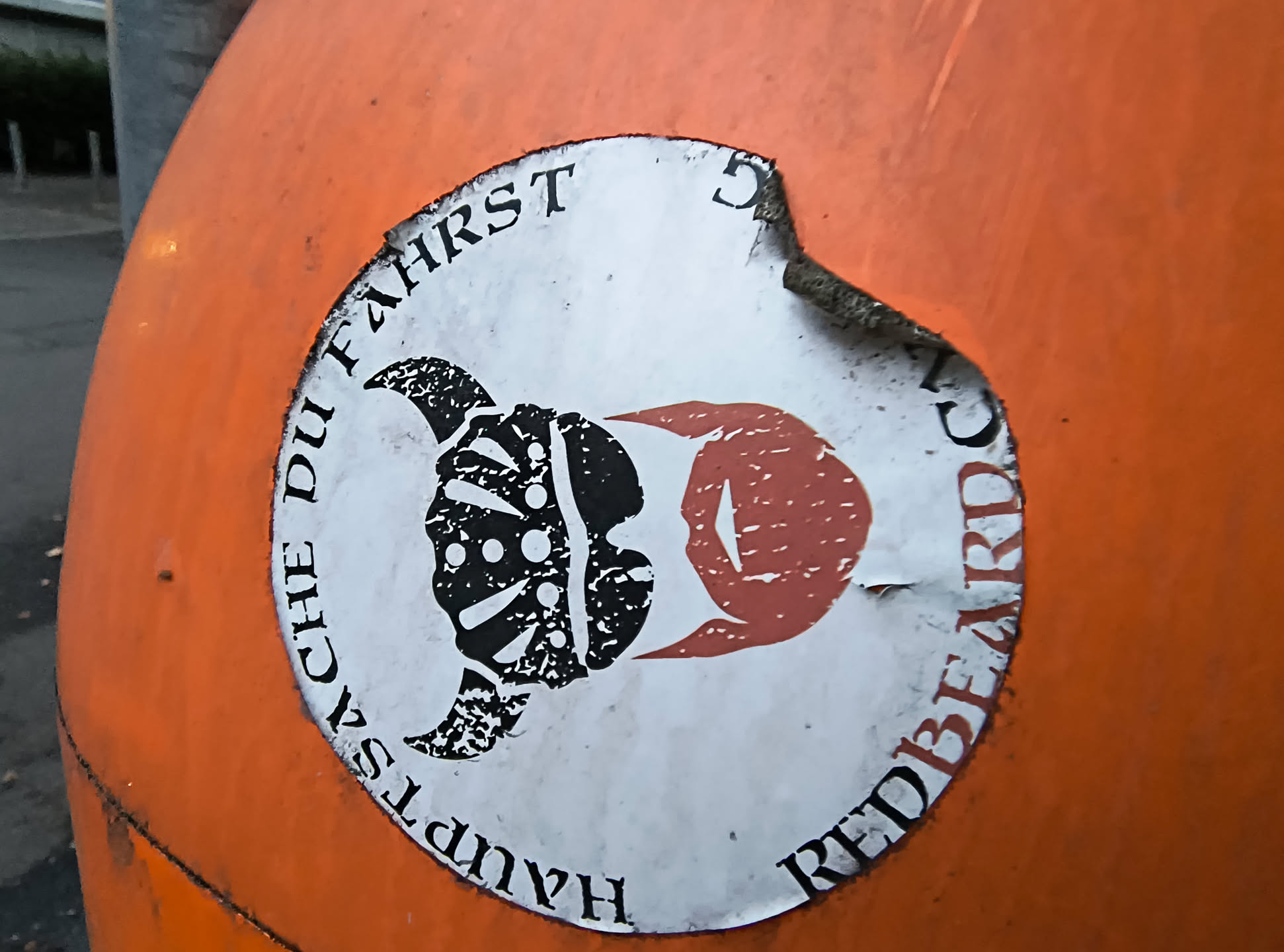 The sticker is circular and predominantly white with black and red graphics. The central image depicts a profile of a head with a horned helmet, rendered in black and white with a textured, almost tribal pattern. To the right of this, a red shape resembling an eye or a flame emanates, also with a textured fill. The text "HAUPTSCHE DU FAHRST" curves around the top left of the circle, and "REDBEARD" curves around the bottom right. The sticker appears to be slightly worn and peeling at the edges, attached to an orange surface.