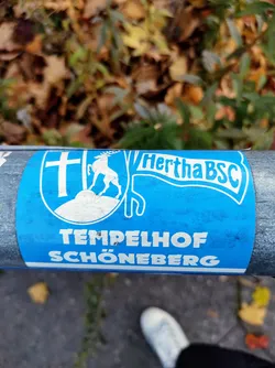 Street sticker A blue sticker featuring the Hertha BSC logo, along with the text 'Tempelhof Schöneberg'. The sticker is affixed to a metal pole outdoors, with autumn leaves visible in the background. 