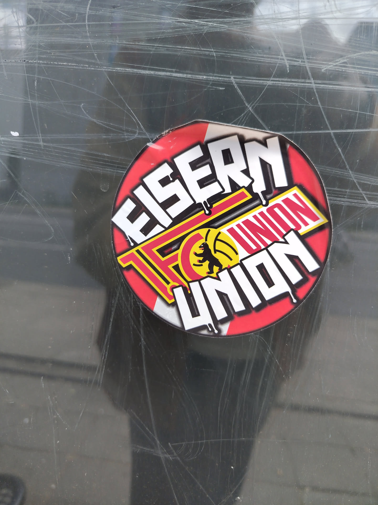 Street sticker A circular sticker predominantly red, featuring large white block letters with black outlines and a 'dripping' effect. The top word reads 'EISERN'. Below it, centered within a yellow rectangular banner outlined in red, is 'FC UNION'. To the left of 'UNION' in this banner, a black silhouette of a bear (representing the Berlin Bear) stands on a yellow circle, set within a red, stylized 'C' or partial circle element. At the bottom of the sticker, the word 'UNION' is repeated in large white dripping letters, similar in style to 'EISERN'. The background behind the text is red, accented with subtle white and grey diagonal lines or a starburst pattern radiating from the center. The design conveys a strong, dedicated, and slightly gritty fan aesthetic.