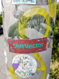 Street sticker A rectangular sticker with the text "ShitVector" written in teal on a maroon background. The sticker has a slightly worn look and is adhered to a gray pole alongside other stickers and graffiti.