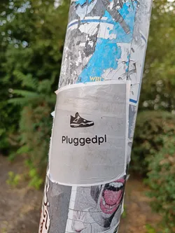 Street sticker A silver sticker with a black silhouette of a sneaker and the text "Pluggedpl" in a sans-serif font. The sticker is affixed to a pole covered in other stickers and graffiti.