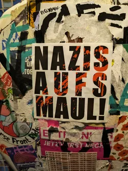 A sticker with the text "Nazis aufs Maul!" in black and orange. The background is white. The text is bold and in all caps. The sticker is attached to a surface with other stickers.